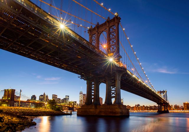 Evening view of Manhattan Bridge with city lights illuminating the skyline in the background. Suitable for promoting travel to New York, design projects highlighting urban architecture, or websites focused on cityscapes and world landmarks.