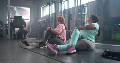Two diverse women stretching in gym performing seated spinal twists with smartwatches