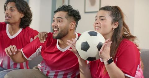 Young fans cheering and holding soccer ball while watching match on couch