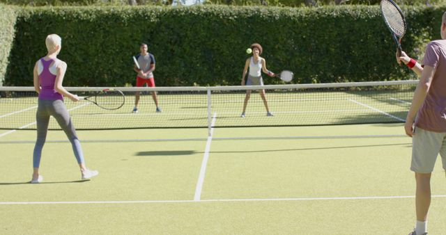 Group of friends playing doubles tennis on bright day. Perfect for themes of teamwork, fitness, sports, diversity, and leisure activities. Suitable for promotional materials, sports magazines, and fitness blogs.