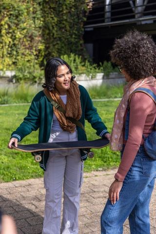 Female friends chatting while holding skateboard on campus walkway with backpacks