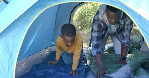 African american father and son preparing sleeping bags inside blue camping tent