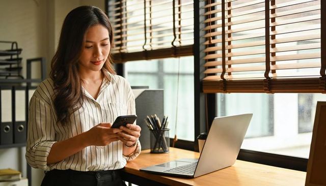 Checking smartphone while working at sunlit modern office desk with laptop and blinds