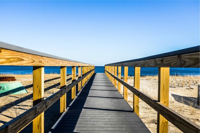 Wooden boardwalk stretches towards ocean with clear blue sky, providing a sense of calm and approach to beach. Perfect for travel websites, beach resort brochures, vacation marketing, and nature blogs highlighting coastal relaxation and serene landscapes.