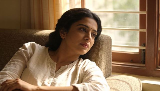 Indian woman gazing thoughtfully on beige sofa wearing white kurta by wooden window