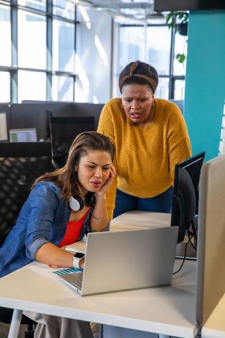 Two diverse professional women collaborating over laptop in modern open-plan office