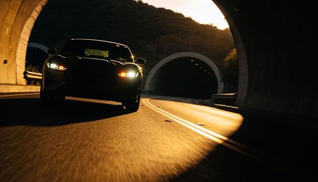 Dark sports car exiting mountain tunnel at golden hour with headlights reflecting on road