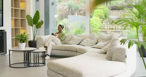 African American woman lounging on curved sofa using smartphone with garden view and indoor plants
