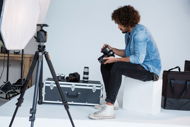 Photographer sitting on a white block, reviewing photos on digital camera in a studio. Surrounded by professional photography equipment including tripod, lighting, and camera gear. Ideal for use in articles about photography, creative professions, studio setups, and professional equipment.