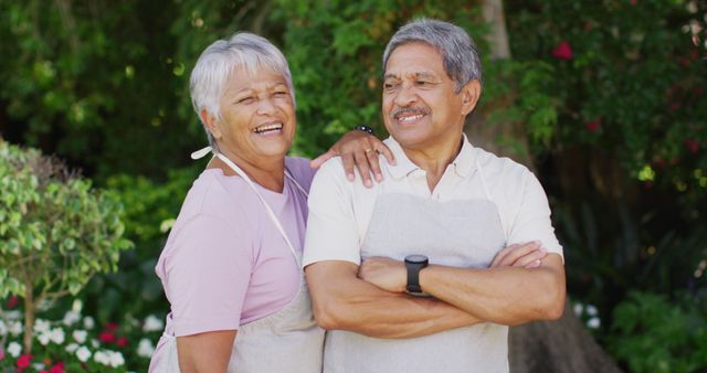 Elderly couple smiling, standing close, and enjoying time together in lush garden. The cheerful seniors are dressed in casual clothing and wearing aprons, implying they might be involved in gardening. Great use for topics related to senior lifestyle, aging gracefully, retirement, outdoor hobbies, and happy relationships.