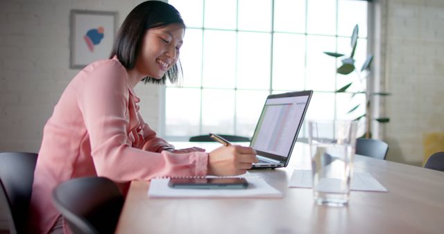 Young businesswoman sitting at desk, working on laptop and taking notes. Bright sunlight from large window gives the office a modern, airy feel. Ideal for commercial use in promotions related to business, professional work environments, telecommuting, or modern office aesthetics.