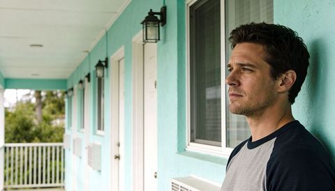 Man leaning against teal motel wall gazing down pastel corridor with doors and ac units
