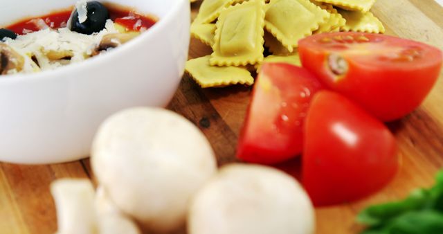 A close-up view of fresh ingredients including tomatoes, mushrooms, and ravioli on a wooden cutting board, with copy space. The focus on the ingredients suggests a preparation for a delicious Italian meal.