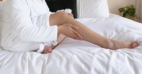 African american woman in white robe applying moisturizer to legs on bed for skincare