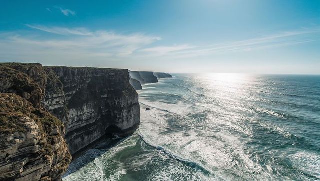 Sunlit sea cliffs towering over foamy surf, layered limestone bluffs and coastal caves