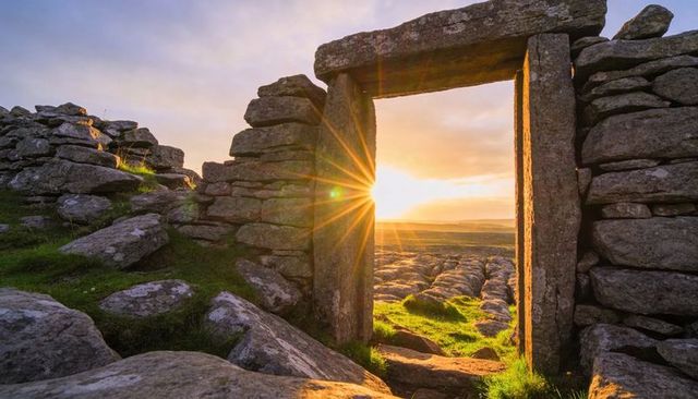 Ancient stone doorway framing sunrise over limestone pavement, sunburst illuminating moor
