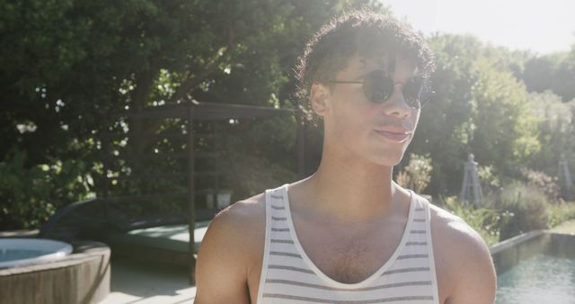 This photo showcases a young man wearing sunglasses and a striped tank top, enjoying a sunny day in an outdoor area with a landscaped garden. Ideal for illustrating time spent outdoors, casual fashion, relaxation, and sunny weather. Suitable for blogs, advertisements, and websites focusing on lifestyle, fashion, and leisure activities.