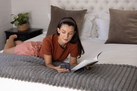 Young woman lying on bed reading paperback and checking smartphone on gray ribbed throw