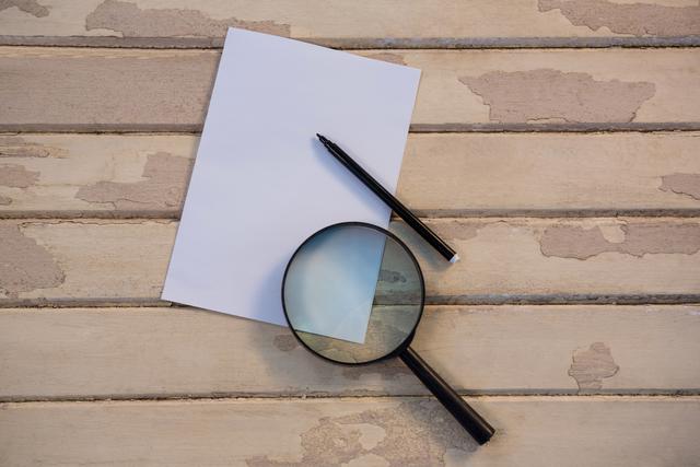 This image shows a magnifying glass, a pen, and a blank sheet of paper on a rustic wooden table. Ideal for themes related to investigation, research, writing, or vintage office supplies. Perfect for use in articles, blogs, or presentations about studying, analyzing documents, or creative writing.