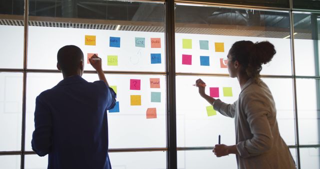 This image shows two colleagues working together, placing colorful sticky notes on an office glass wall. Ideal for use in business, teamwork, idea generation, project planning, and innovation-related contexts. Suitable for illustrating modern office environments, collaborative work processes, and strategic meetings.