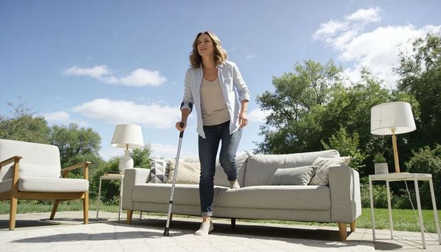 Woman using forearm crutch balancing on outdoor sofa on sunlit patio with cushions and green lawn
