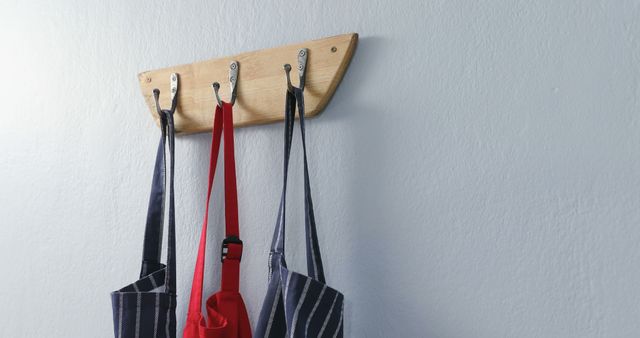 Colorful aprons, including red and striped styles, hang on wooden wall hooks. This setting can be used to showcase kitchen organization ideas, home decor tips, or articles on cooking and kitchen setups. It appeals to audiences interested in neat and organized living spaces.