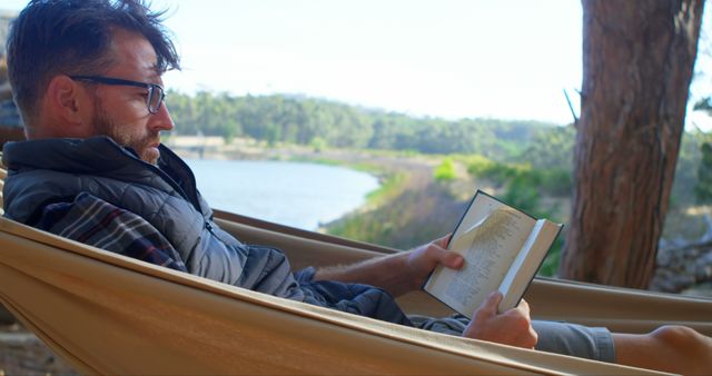 Man relaxing in a hammock while reading a book, set against the backdrop of a serene lake and lush greenery. Ideal for promoting outdoor leisure activities, reading retreats, and emphasizing the importance of relaxation amidst nature. Suitable for use in travel brochures, articles on outdoor lifestyle, and wellness blogs.