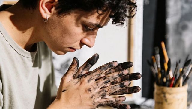Young artist rubbing ink-stained hands while inspecting sketch in sunlit studio workspace