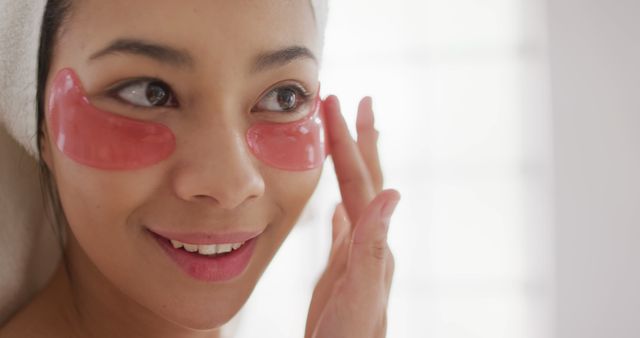 The scene shows an Asian woman with a towel wrapped around her head, using pink under-eye gel patches. She is smiling softly while gently touching her cheek, indicating a skincare routine. This content can be used for beauty and skincare articles, wellness blogs, self-care tutorials, and spa advertisements.