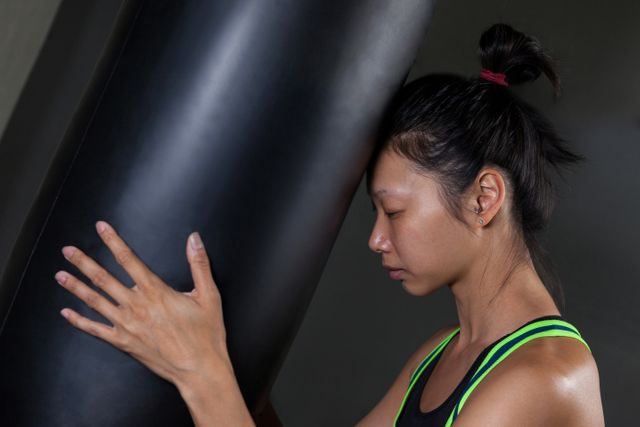 Female boxer leaning on a punching bag, showing exhaustion and determination after an intense workout in a fitness studio. Ideal for use in fitness, sports, and motivation-related content, highlighting themes of perseverance, strength, and dedication.