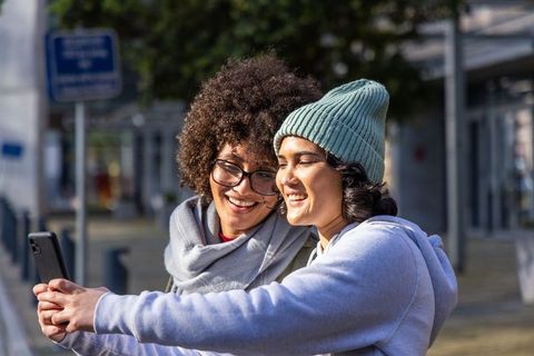 Smiling friends taking selfie on sunny urban sidewalk wearing beanie, scarf, eyeglasses