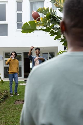 Multiethnic friends throwing football on suburban lawn by modern white house, casual outdoor play