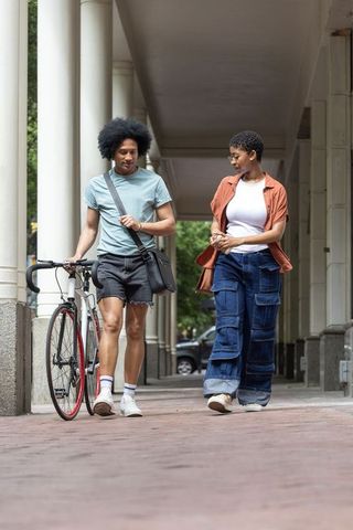 African american couple walking with bicycle along urban colonnade chatting during commute