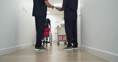 Nurses passing metal tray in hospital corridor with wheelchair and supply cart