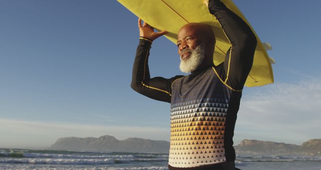 This image shows a senior man holding a yellow surfboard over his head while standing on a sunny beach with mountains in the background. Suitable for promoting active lifestyles among seniors, surf culture, water sports, beach vacations, and healthy living during retirement.