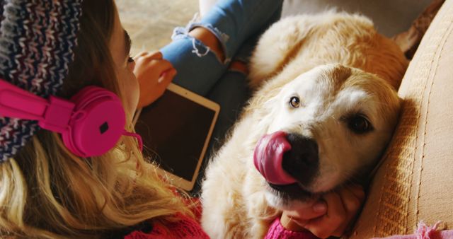 Image showing a young woman lounging at home, using a tablet, and wearing pink headphones while her Golden Retriever relaxes next to her. A sense of comfort, relaxation, and companionship is evident. Ideal for advertising home and lifestyle products, promoting pet care brands, and illustrating themes of bonding and technology in everyday life.