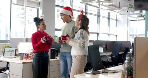 Diverse coworkers exchanging holiday gifts wearing santa hat and reindeer headband in open office