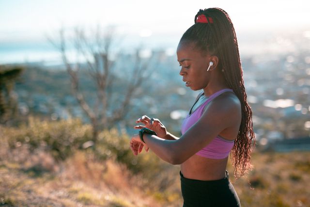 This image depicts a fit African American woman wearing sportswear and earphones, checking her smartwatch during an outdoor workout in the countryside. Ideal for use in articles or advertisements related to fitness, health, technology in sports, outdoor activities, and active lifestyles.