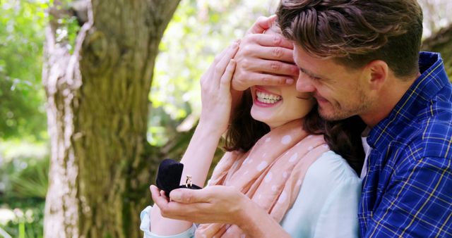 A young Caucasian man surprises a woman by covering her eyes and presenting an engagement ring, with copy space. Their joyful expressions and the outdoor setting suggest a romantic and significant life event.
