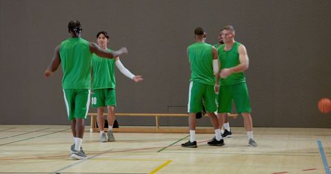 Diverse male basketball team warming up and greeting in indoor gym, teamwork and sportsmanship