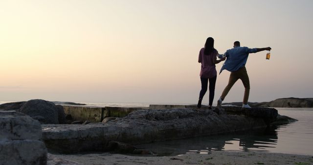 Biracial couple balancing on rocks by sea, holding hands. Both have dark hair, the woman wearing pink top, the man in a blue shirt