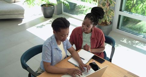 African mentor guiding teen on laptop in bright home workspace with plants and sunlight