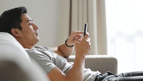 Reclining asian man scrolling smartphone on gray sofa, relaxed minimalist home lifestyle