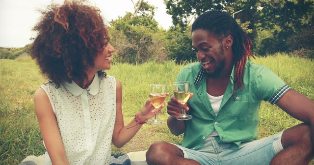 A young African American couple enjoys a romantic picnic, toasting with glasses of wine, with copy space. Their cheerful interaction in a natural setting suggests a moment of celebration or a casual outdoor date.