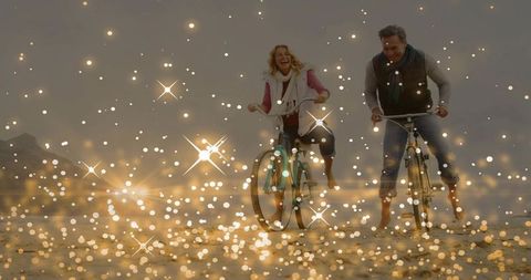 Mature couple riding bicycles on wet beach at dawn with golden bokeh sparkle overlay