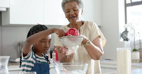 Senior woman guiding young girl sifting flour while baking together in modern kitchen