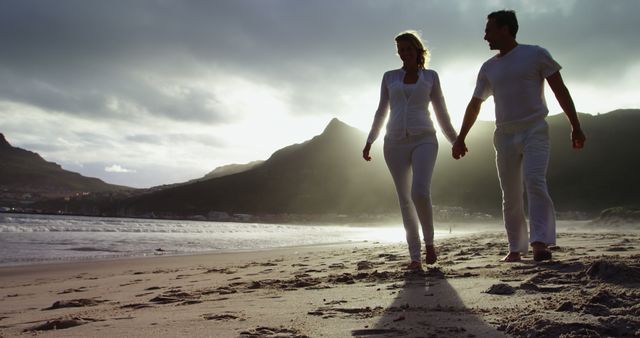 Romantic couple walking hand in hand along a tranquil beach with mountains in the background and sunset casting a beautiful glow. Perfect for promoting destinations, travel brochures, romantic getaways, wellness retreats, and lifestyle blogs.
