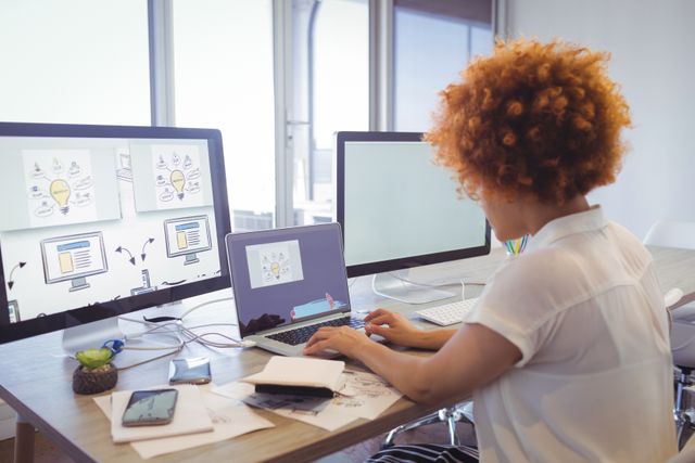 Businesswoman concentrating on work at her desk in a modern office with multiple monitors. Ideal for use in professional, business, and technology related content. Depicts themes of productivity, multitasking, and modern work environments.