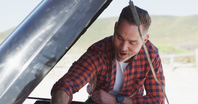 Man wearing a plaid shirt, inspecting or fixing a car engine outdoors on a sunny day. Ideal for content related to automotive repair, do-it-yourself maintenance, mechanical work, travel preparedness, and problem-solving skills.