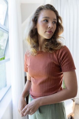 Young woman standing by window gazing through curtains in rust top and belted green skirt